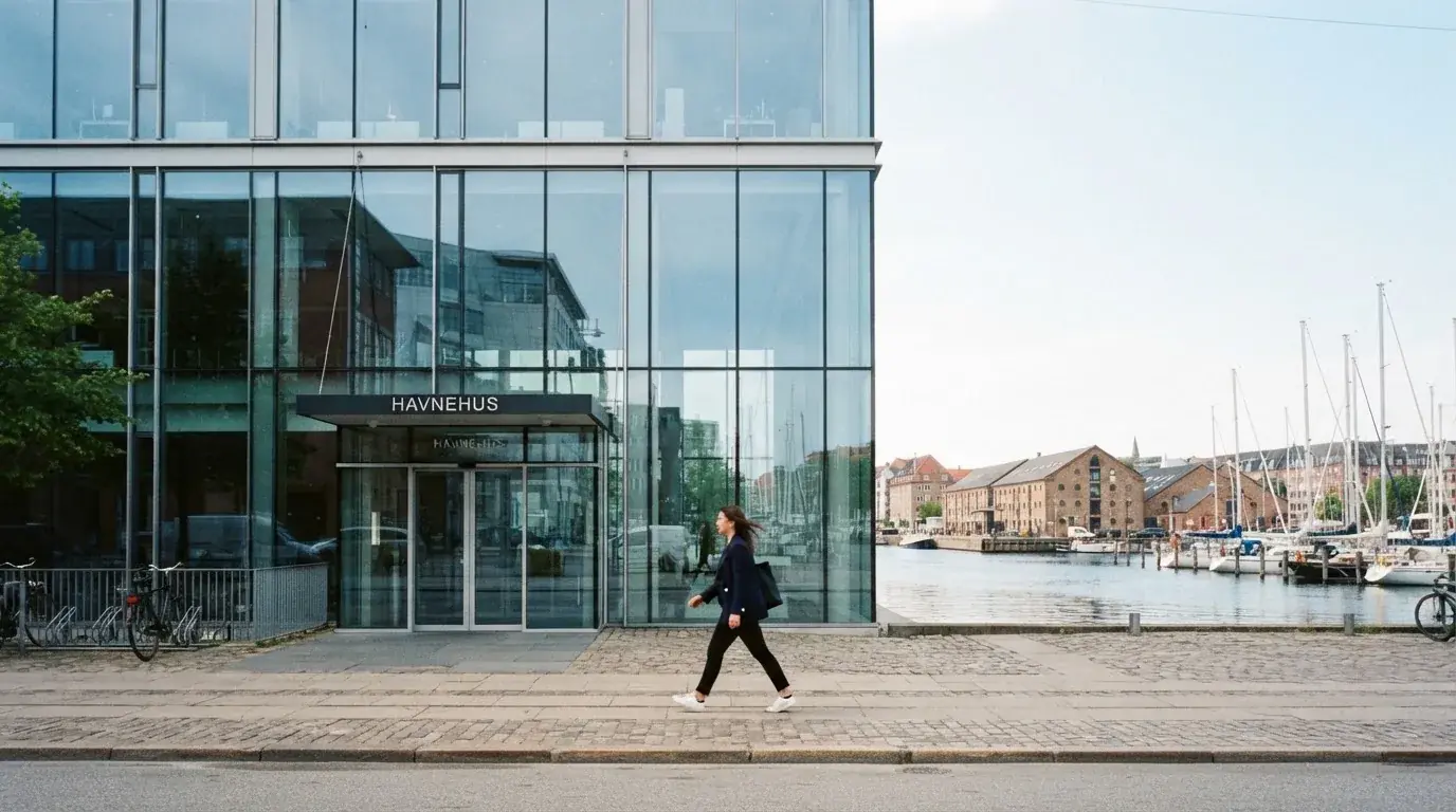 En moderne kontorbygning med glasfacade ved Københavns havn i klart dagslys, med en medarbejder i blazer, der går forbi.
